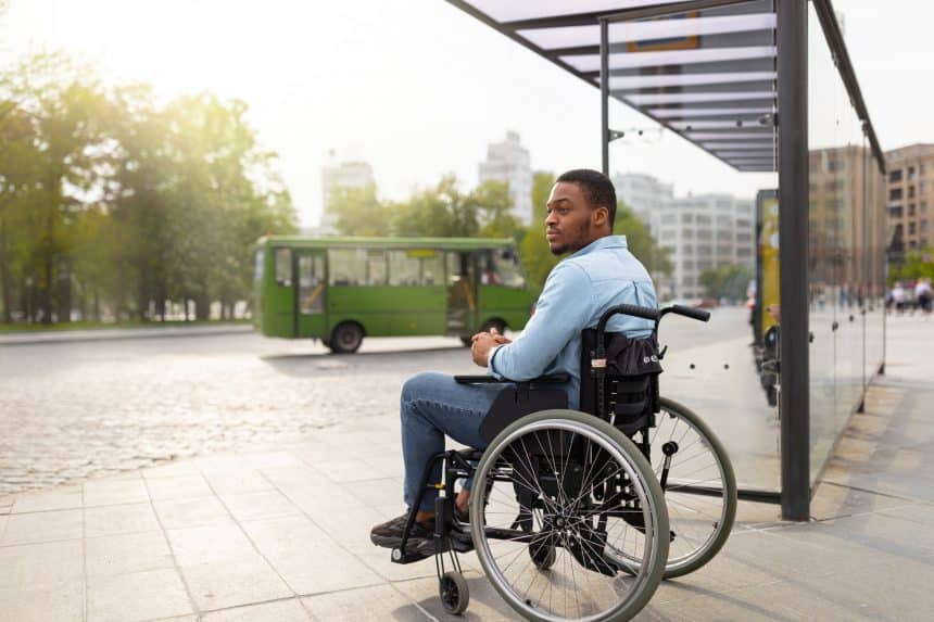 Young disabled black man in wheelchair waiting for public transport on bus stop, having difficulty